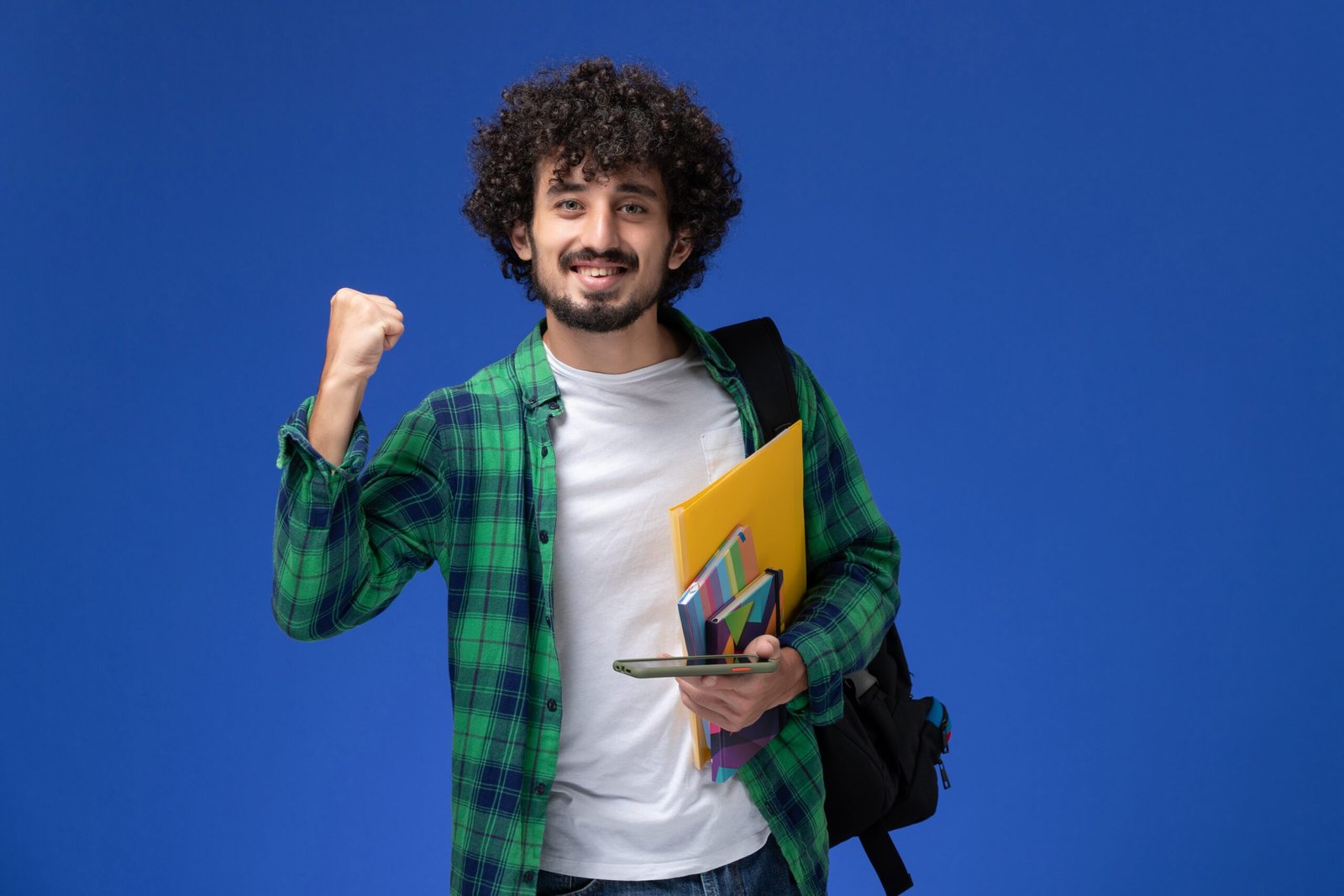 front view male student wearing black backpack holding copybook files using his phone light blue wall