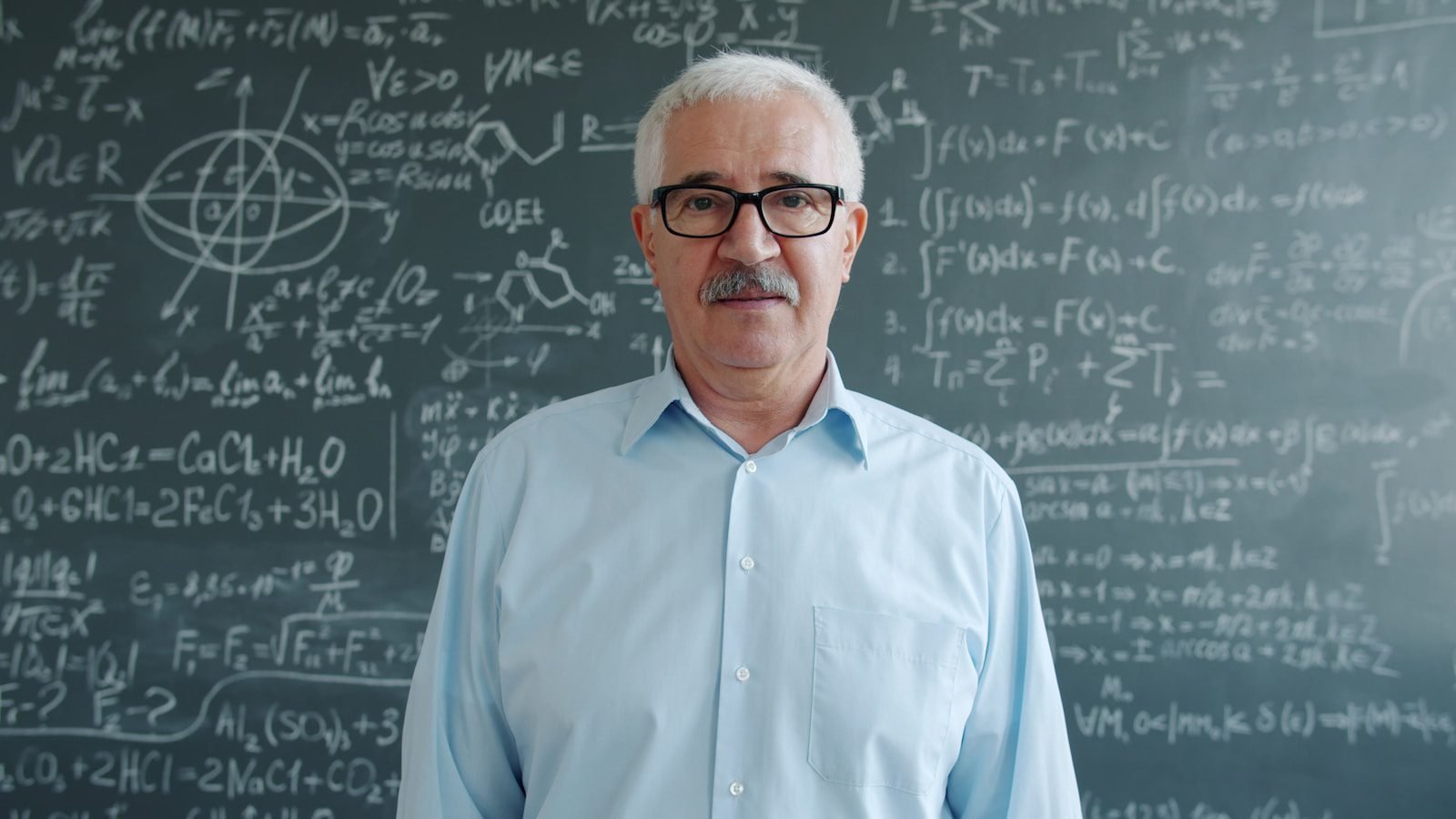 Elderly man in glasses stands before chalkboard with math equations.